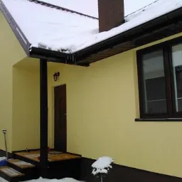 Żółty dom jednorodzinny z ciemnobrązowymi oknami i dachem pokrytym śniegiem, widoczne schody wejściowe i łopata oparta o ścianę.