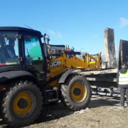Żółta koparko-ładowarka JCB transportuje betonowe płyty na przyczepie, pracownik w kamizelce odblaskowej kontroluje załadunek, w tle widać pryzmę ziemi.