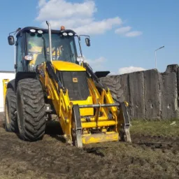 Żółta koparko-ładowarka JCB na placu budowy, widok z przodu, z oponami lekko zabrudzonymi błotem, w tle betonowy mur i budynek magazynowy.