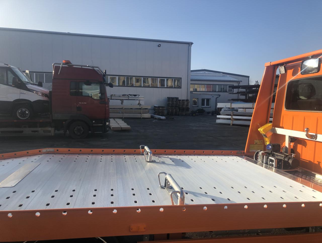 Pomarańczowa laweta z załadowanym białym vanem na tle szarego budynku przemysłowego w słoneczny dzień.