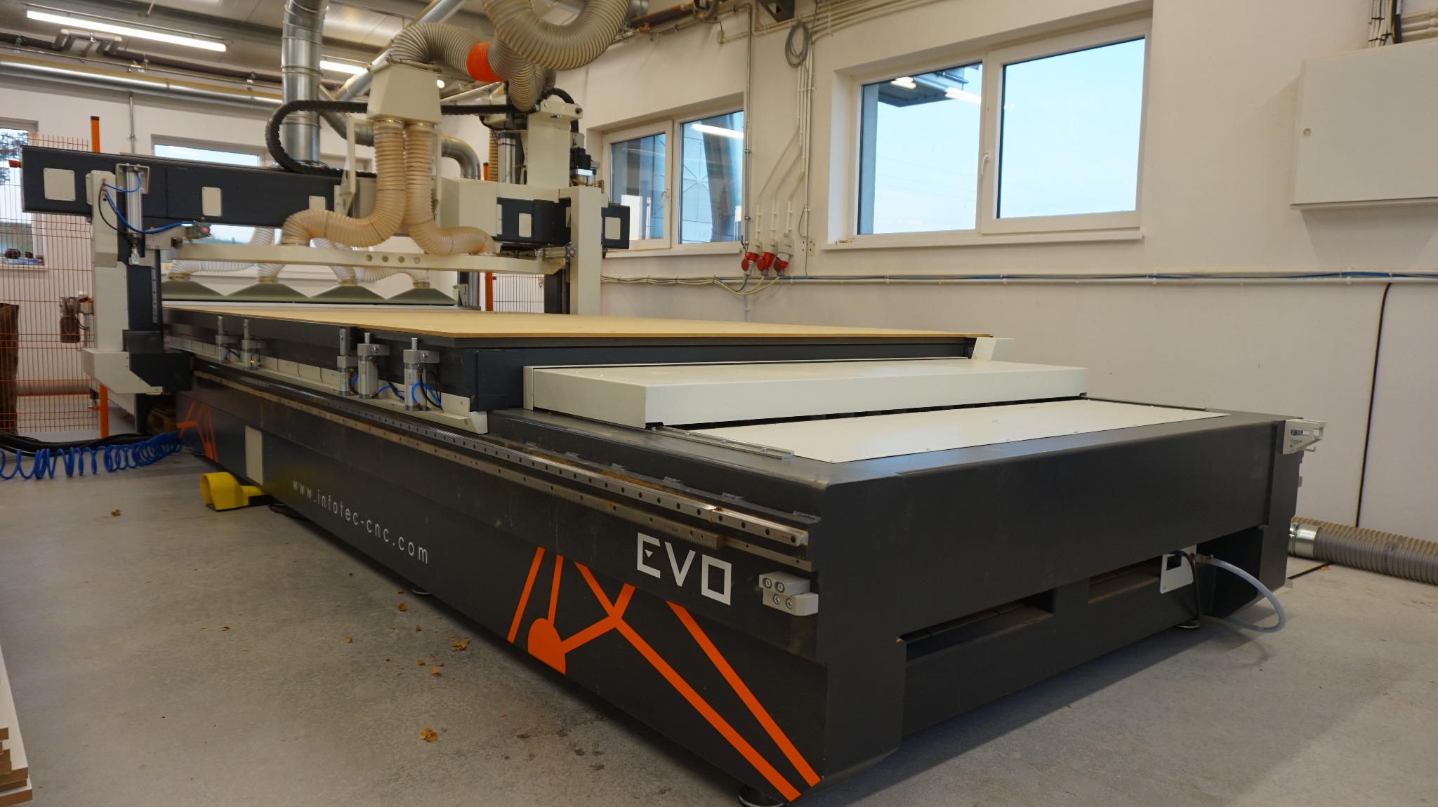 Duża frezarka CNC EVO w hali produkcyjnej przygotowana do obróbki płyty drewnianej, widoczne przewody wentylacyjne i okna w tle.