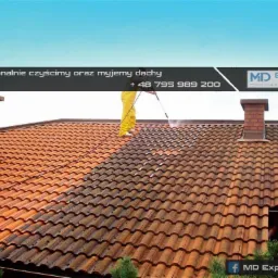 Pracownik w żółtym kombinezonie czyści dach z czerwonej dachówki w Bełchatowie za pomocą myjki ciśnieniowej; widoczna różnica w kolorze pomiędzy czystą i brudną częścią dachu, komin z cegły w tle.