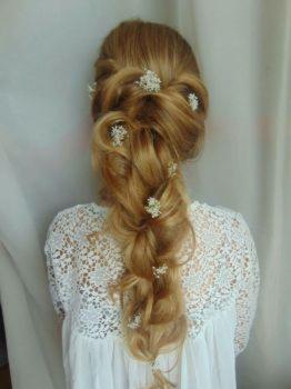 Uczesanie ślubne z wplecionymi drobnymi białymi kwiatami, włosy blond upięte w luźny warkocz od tyłu, biała koronkowa bluzka.