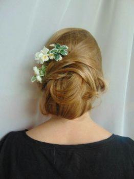 Upięte blond włosy z wpiętymi białymi kwiatami z zielonymi liśćmi, widok z tyłu, na tle białej, delikatnie pofałdowanej tkaniny.