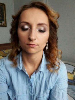 Portret kobiety z delikatnym makijażem oczu w srebrnych odcieniach i upiętymi, rudawymi włosami, pozującej w pomieszczeniu z widocznym łóżkiem i obrazem w tle.