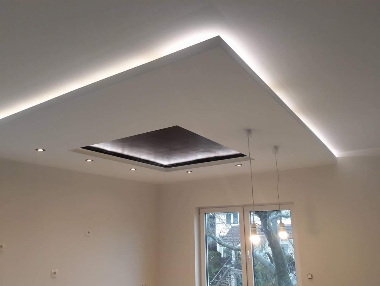 Nowoczesny sufit podwieszany z oświetleniem LED wzdłuż krawędzi i centralnym kwadratem oświetlonym punktowo, widok na okno i drzewo na zewnątrz.