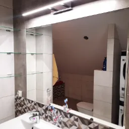 Duże lustro w łazience z widocznymi szklanymi półkami i odbiciem wnętrza, w tym toalety i pralki.