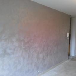 Wykończenie wnętrz marcos - Ściana w pokoju pokryta dekoracyjną, metaliczną farbą w odcieniach szarości i beżu, z widoczną fakturą imitującą chmurki lub marmurkowanie, częściowo oświetlona przez światło słoneczne