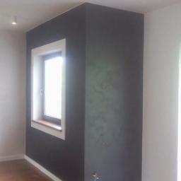 Wykończenie wnętrz marcos - Pomieszczenie po świeżo wykonanych pracach: ściana pomalowana na ciemny kolor z widoczną fakturą, biała rama okienna i listwy przypodłogowe, drewniana podłoga, widoczne punkty świetlne na suficie.