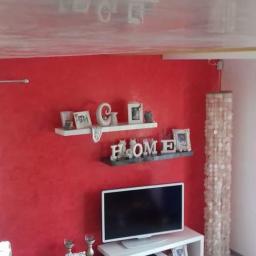 Wykończenie wnętrz marcos - Czerwona ściana z dekoracyjną fakturą, biały sufit z połyskiem, dwie półki z dekoracjami, biała szafka RTV z telewizorem, oraz lampa z białych krążków.