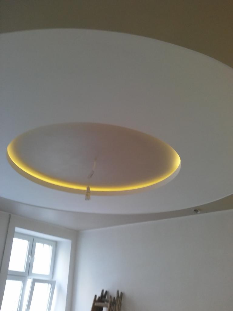 Okrągły sufit podwieszany z oświetleniem LED, widoczny fragment okna i ściany, dolny kadr z drabinką malarską.
