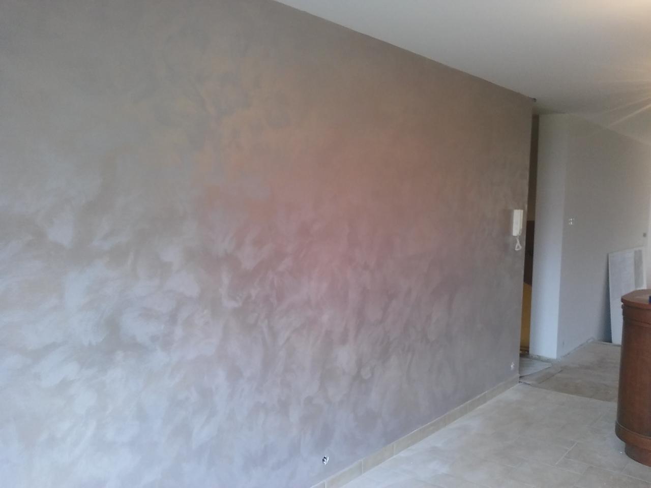 Ściana w pokoju pokryta dekoracyjną, metaliczną farbą w odcieniach szarości i beżu, z widoczną fakturą imitującą chmurki lub marmurkowanie, częściowo oświetlona przez światło słoneczne