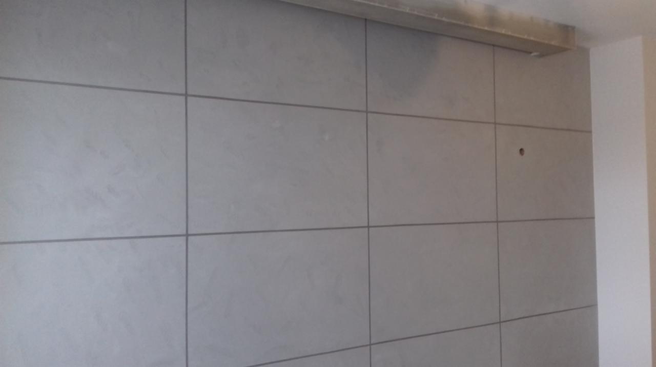 Ściana pokryta dużymi, szarymi płytami z widocznymi spoinami; widoczny fragment sufitu z elementem konstrukcyjnym oraz narożnik ściany.