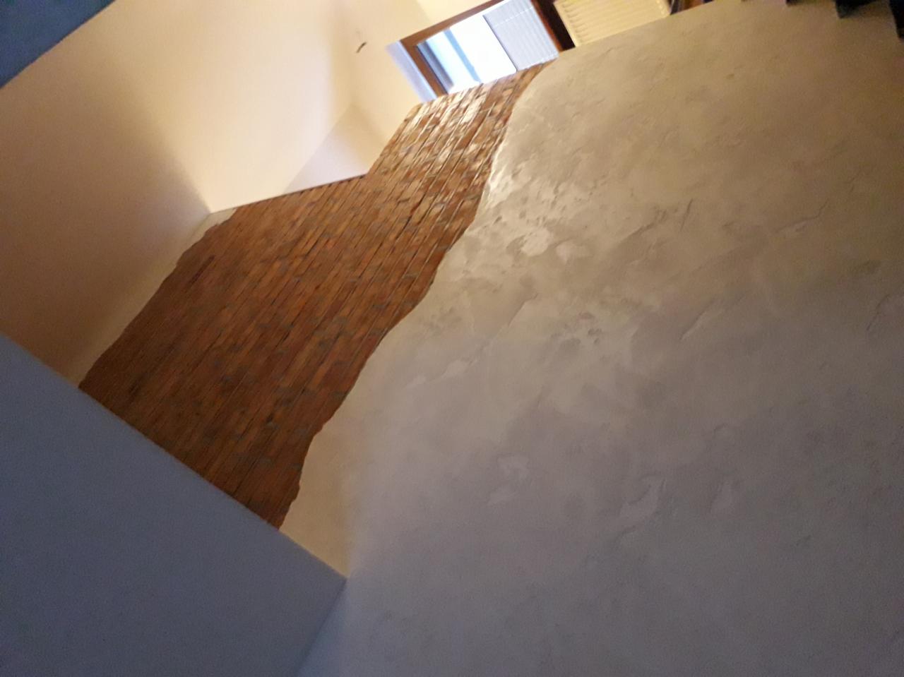 Wnętrze z dekoracyjną ścianą: fragment muru z czerwonej cegły sąsiaduje z gładzią w odcieniach szarości, widok z dołu w kierunku okna.