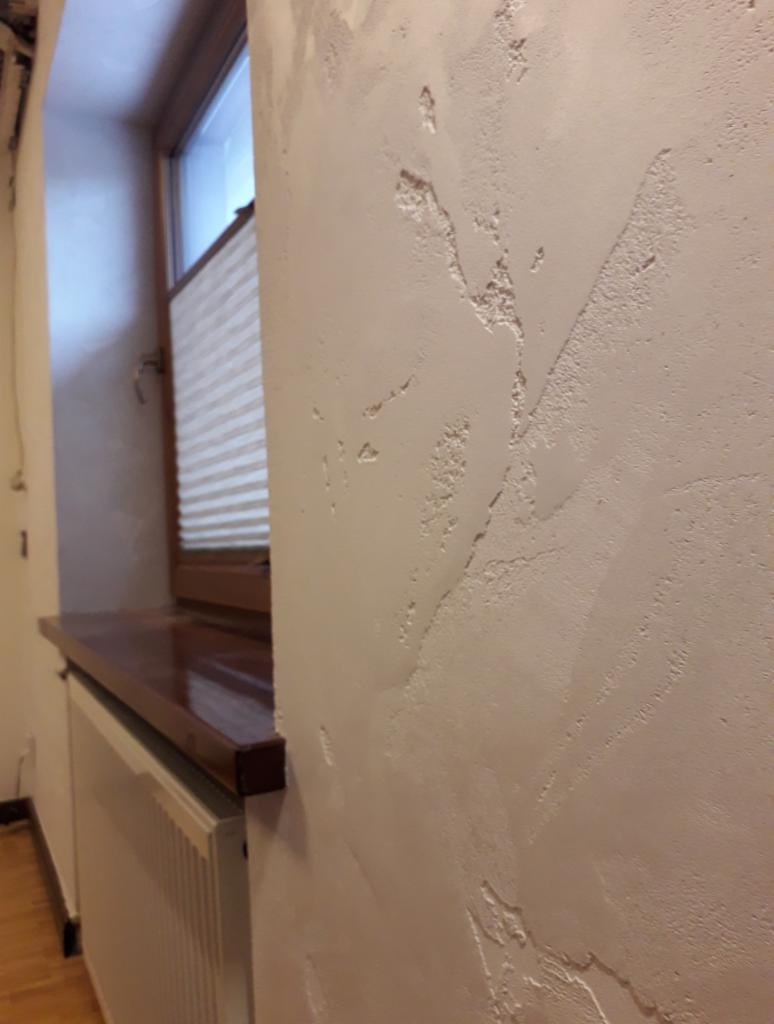 Ściana z dekoracyjną strukturą przypominającą naturalny kamień, obok okna z drewnianą ramą i plisowaną roletą, nad grzejnikiem z drewnianym blatem.