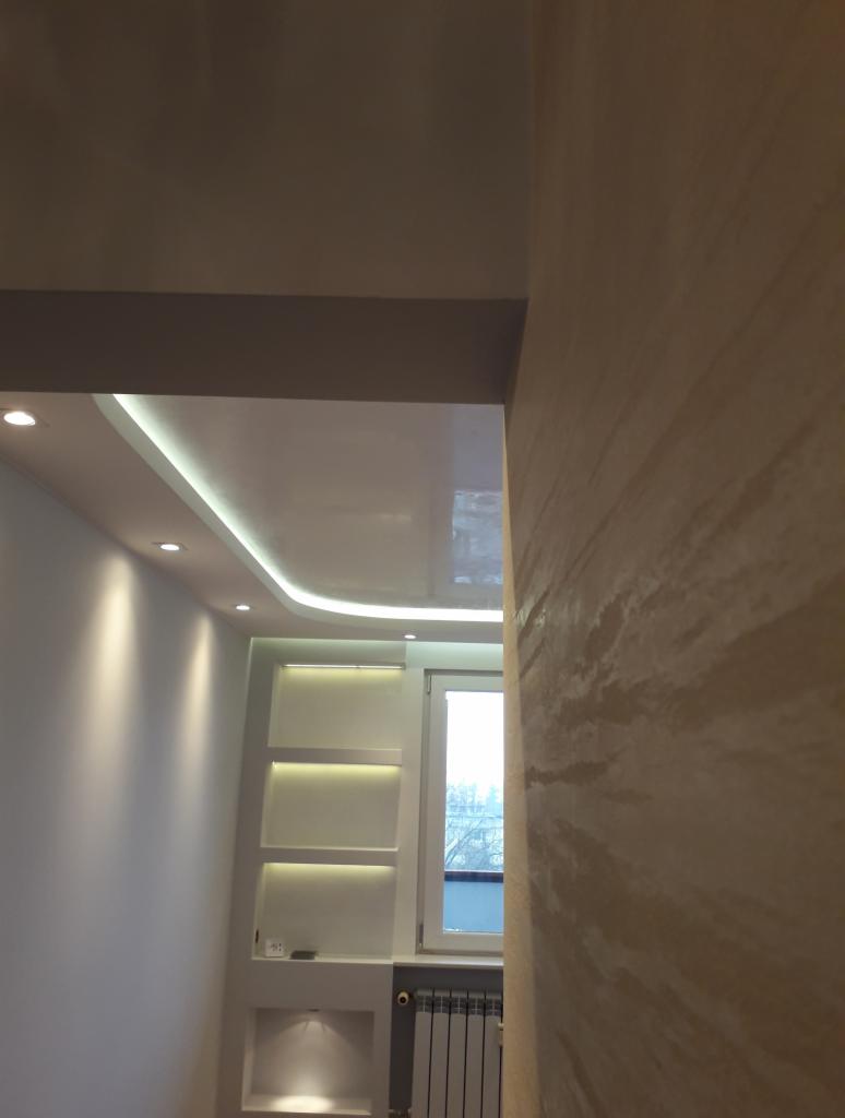 Wykończona ściana z efektem dekoracyjnym, widoczny podwieszany sufit z oświetleniem LED oraz wnęka z półkami i oświetleniem punktowym, obok okno z grzejnikiem.
