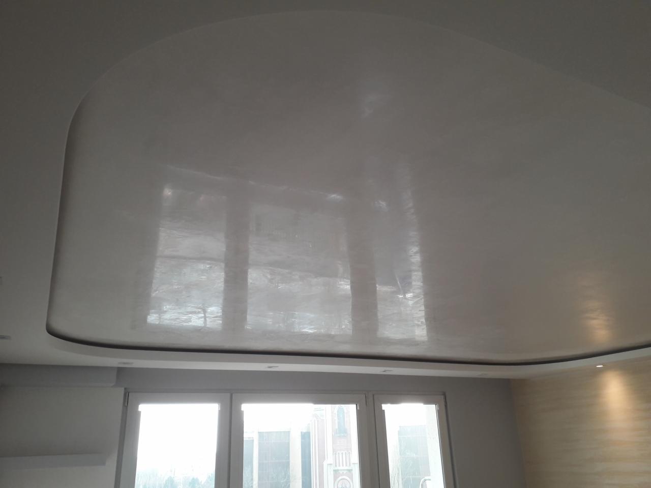 Gładka, biała, zaokrąglona powierzchnia sufitu z widocznym odbiciem okien, wykończona z subtelnym oświetleniem punktowym i panelami ściennymi w tle.