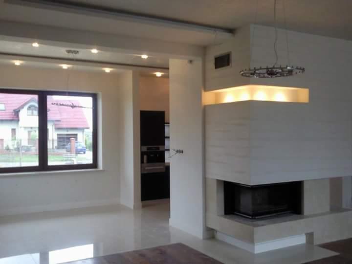 Jasne wnętrze z kominkiem obudowanym beżowym kamieniem, nowoczesnym oświetleniem i widokiem na dom z czerwonym dachem przez okno.