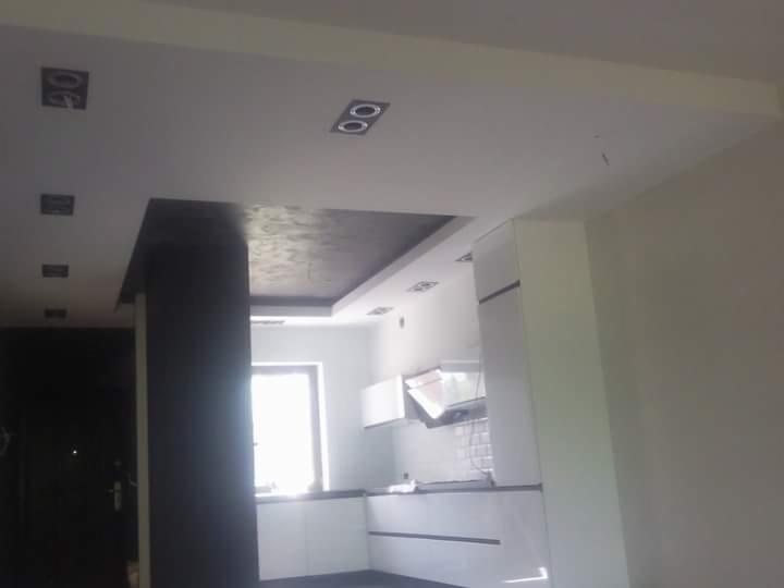 Wykończone wnętrze z ciemną wnęką sufitową i kolumną, białe ściany, oświetlenie punktowe, widoczny fragment kuchni z białymi szafkami.