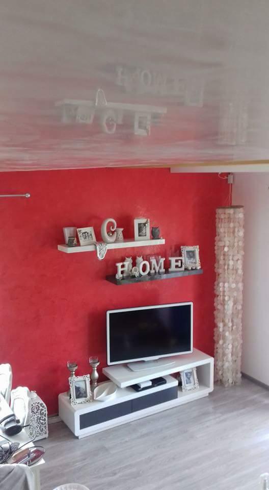 Czerwona ściana z dekoracyjną fakturą, biały sufit z połyskiem, dwie półki z dekoracjami, biała szafka RTV z telewizorem, oraz lampa z białych krążków.