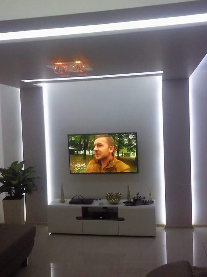 Nowoczesny salon z podświetlanymi pionowymi i poziomymi liniami LED, telewizorem na ścianie i białą szafką RTV, uzupełniony rośliną doniczkową w rogu.