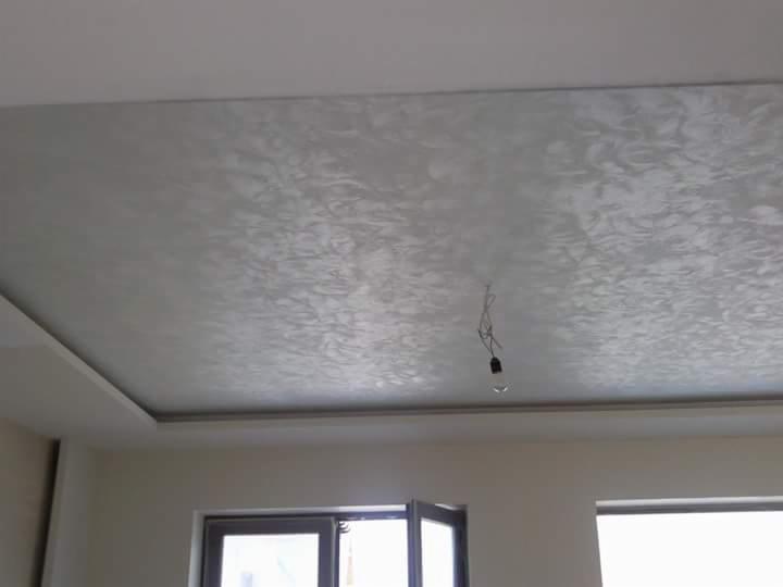 Wykończony sufit z dekoracyjną, srebrzystą fakturą, widoczne okna i wystający fragment sufitu z boku, wisząca żarówka na kablu.