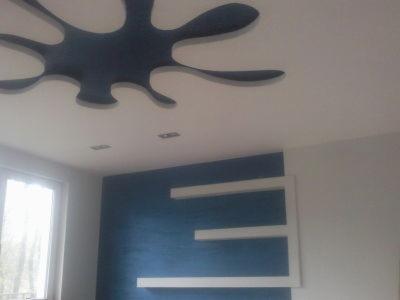 Niebieska ściana z trzema białymi półkami w kształcie litery E, biały sufit z czarną dekoracją w kształcie kleksa i oknem z widokiem na drzewa.