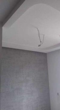 Świeżo wykończone wnętrze w Łodzi z szarą ścianą o fakturze betonu i białym, podwieszanym sufitem z zaokrąglonymi krawędziami, widoczne wystające kable elektryczne.