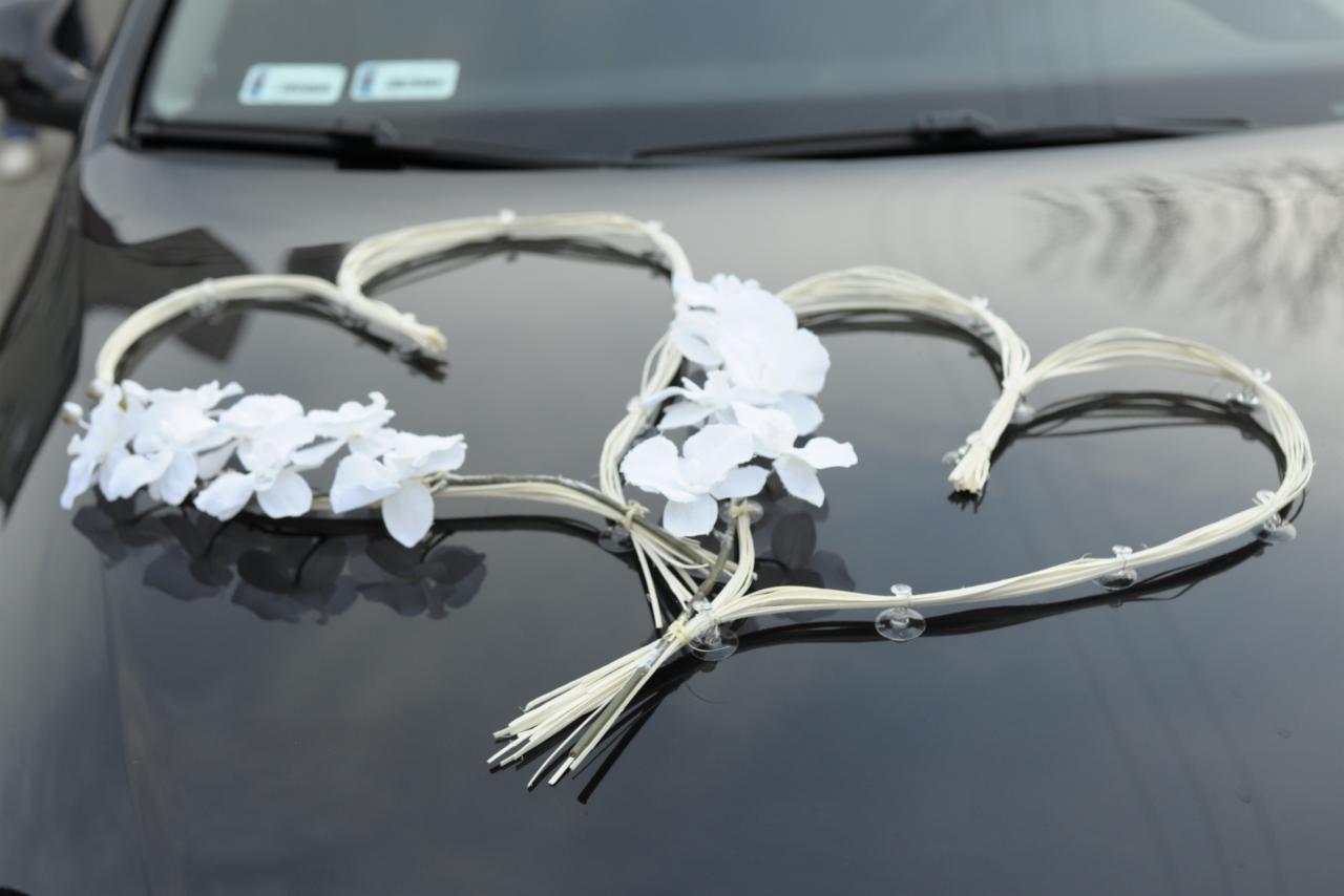 Dekoracja ślubna na samochodzie: dwa serca z białych kwiatów i rafii przymocowane przyssawkami do lakierowanej maski.