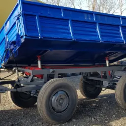 Błyszcząca, niebieska przyczepa wywrotka z czerwonymi siłownikami hydraulicznymi podniesiona do góry, stojąca na żwirowym podłożu.