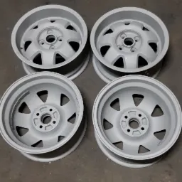 Cztery odnowione felgi samochodowe, pokryte świeżą warstwą szarego podkładu, leżące na betonowej posadzce warsztatu.