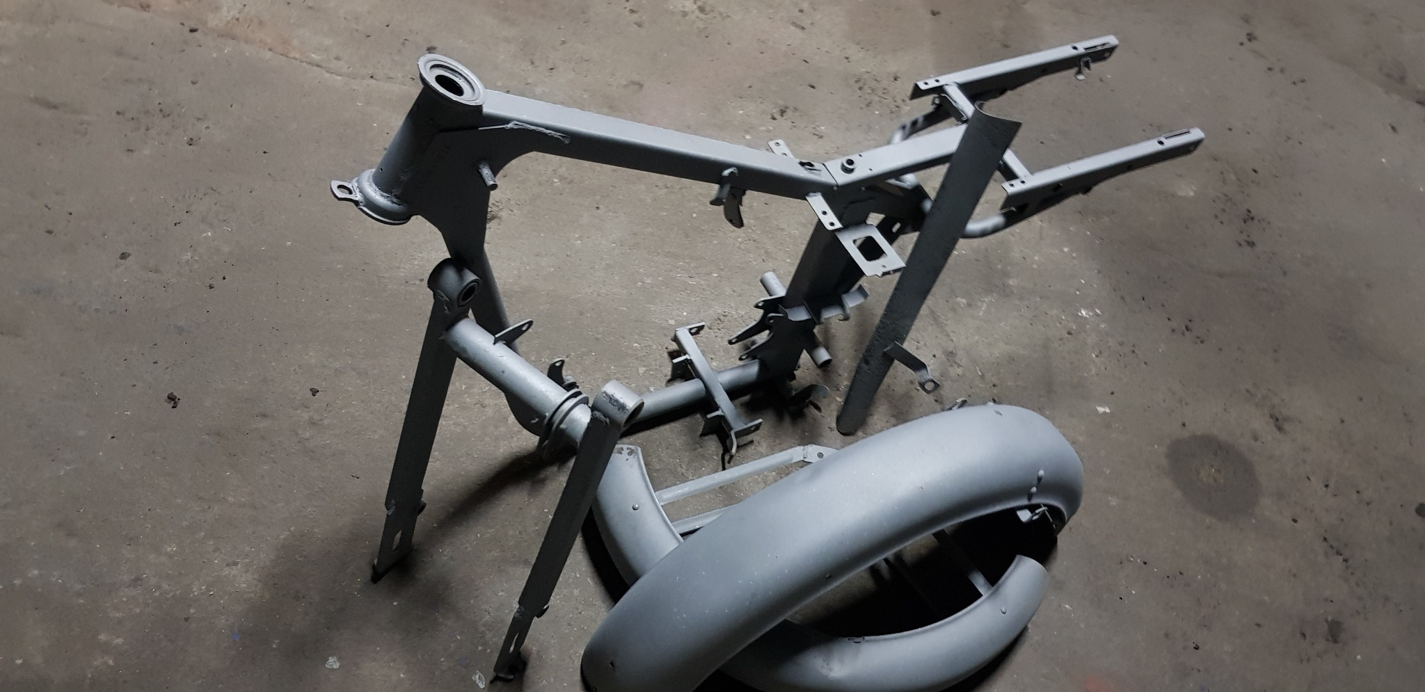 Szare elementy ramy motocyklowej i błotnik po obróbce, leżące na betonowej podłodze w warsztacie.