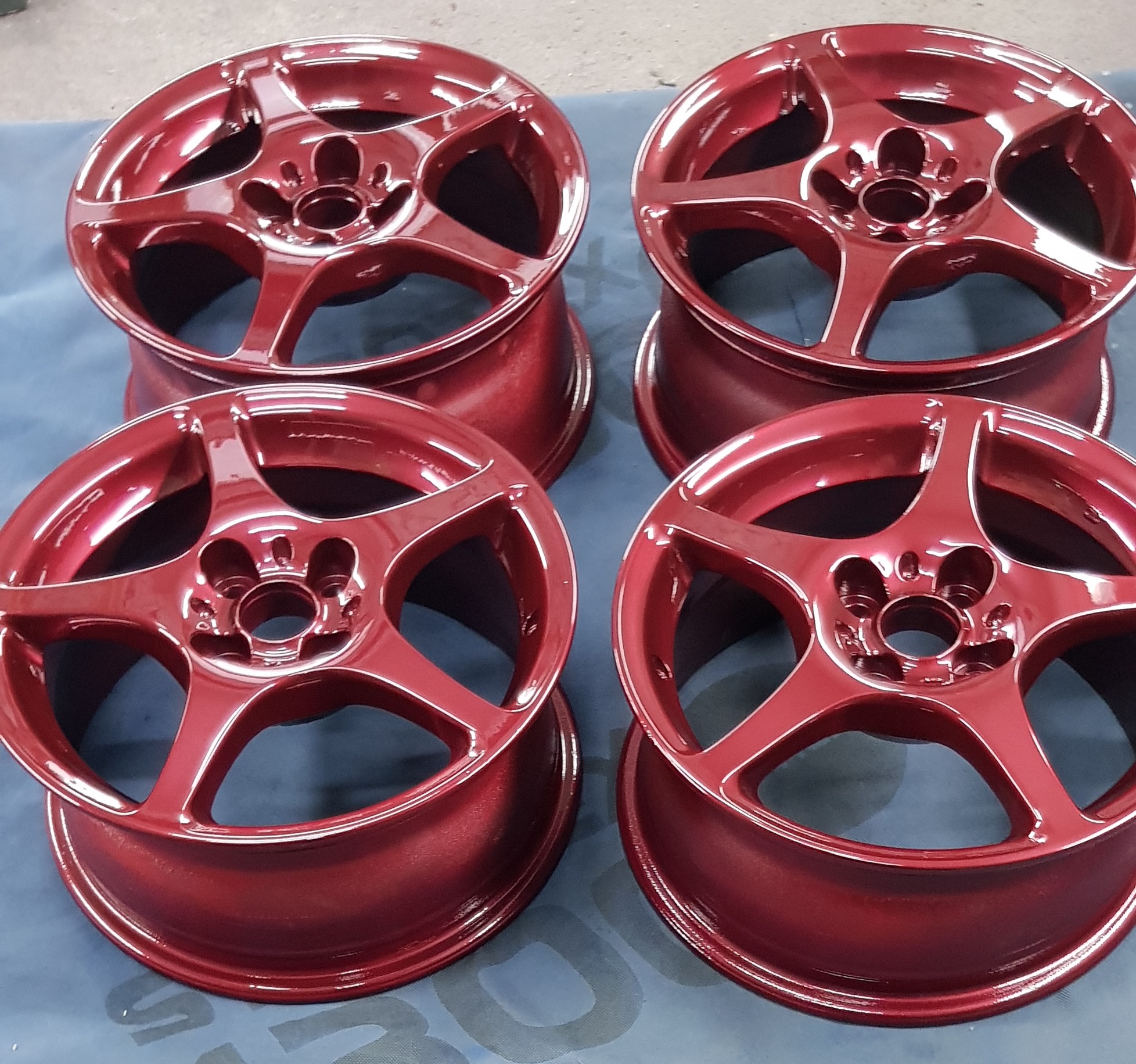 Cztery felgi aluminiowe po renowacji, pomalowane na metaliczny odcień czerwieni, ułożone parami na niebieskim tle.