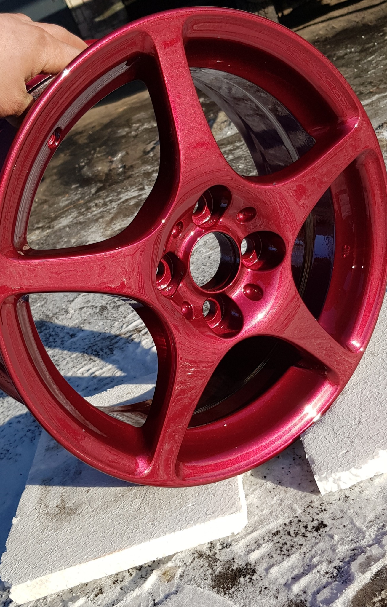 Błyszcząca, bordowa felga samochodowa z pięcioma ramionami, świeżo po renowacji, leży na białym styropianie na tle śniegu.