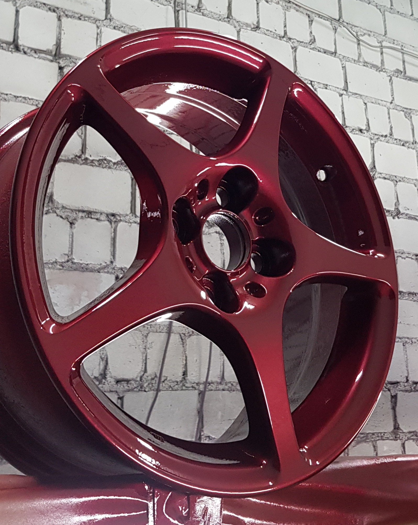 Błyszcząca, bordowa felga aluminiowa z pięcioma ramionami, po malowaniu proszkowym, na tle białej ściany z cegieł.