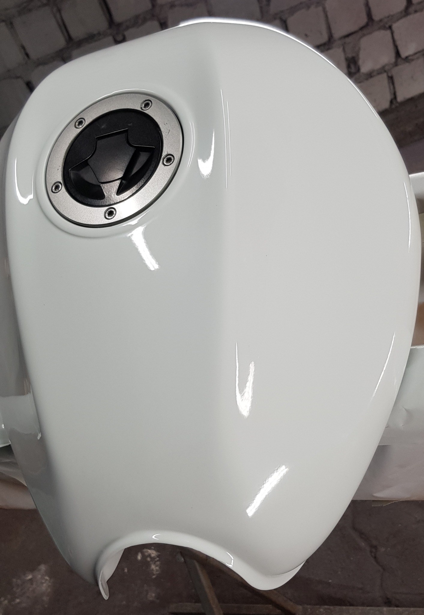 Biały, świeżo polakierowany zbiornik paliwa motocyklowego z widocznym wlewem paliwa, umieszczony w warsztacie z ceglaną ścianą w tle.