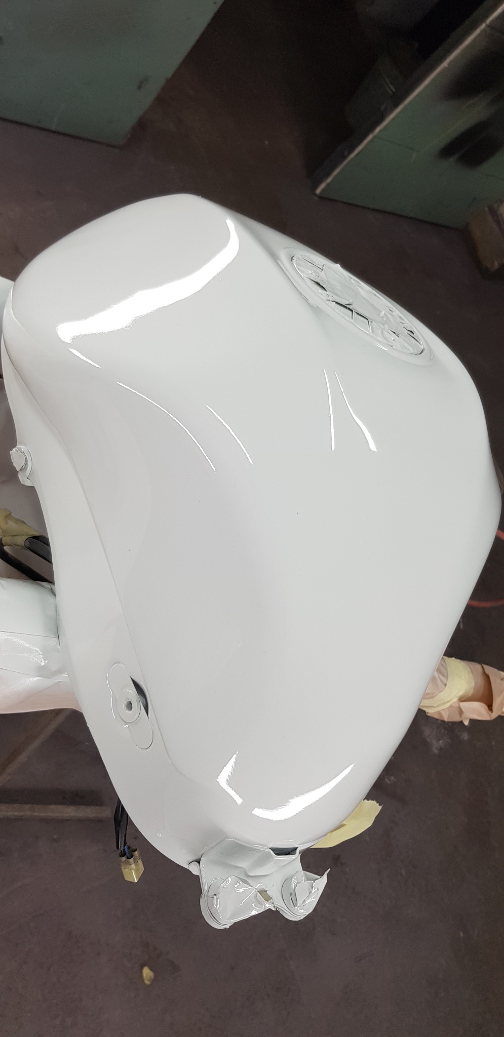 Błyszczący, biały bak motocyklowy po lakierowaniu, zabezpieczony taśmą malarską w dolnej części, widoczne odbicia światła na powierzchni.