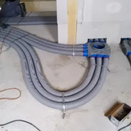 Szare, karbowane rury instalacyjne prowadzone w ścianie, zabezpieczone metalowymi obejmami, widoczne puszki rozdzielcze z niebieskimi elementami i fragment izolacji z wełny mineralnej w surowym...