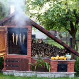 Murowana wędzarnia z cegły z otwartymi drzwiczkami, wewnątrz wędzone ryby, obok stos drewna, dynie i ozdobny dzbanek na tle zielonego ogrodu.