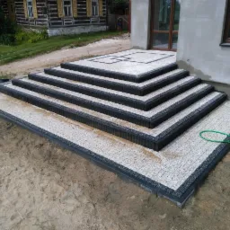 Nowo ułożone schody zewnętrzne z kostki brukowej w odcieniach szarości i czerni, prowadzące do wejścia domu, z widocznym fragmentem trawnika i drewnianego budynku w tle.