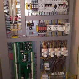 Otwarta szafa sterownicza z widocznymi modułami elektronicznymi, przekaźnikami, bezpiecznikami, listwami zaciskowymi i okablowaniem. Widoczna płytka sterująca z zielonym laminatem i licznymi...