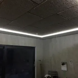 Panelowy sufit z oświetleniem LED wzdłuż krawędzi, widoczne przewody wystające z sufitu, ściany w odcieniach szarości, okno balkonowe.