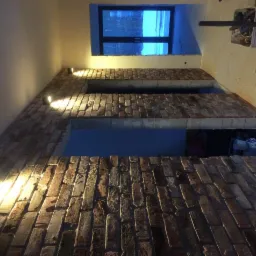 Ceglana ściana z wnękami oświetlona punktowymi lampami, widok z dołu, w tle okno z widokiem na zewnątrz i narzędzia budowlane.