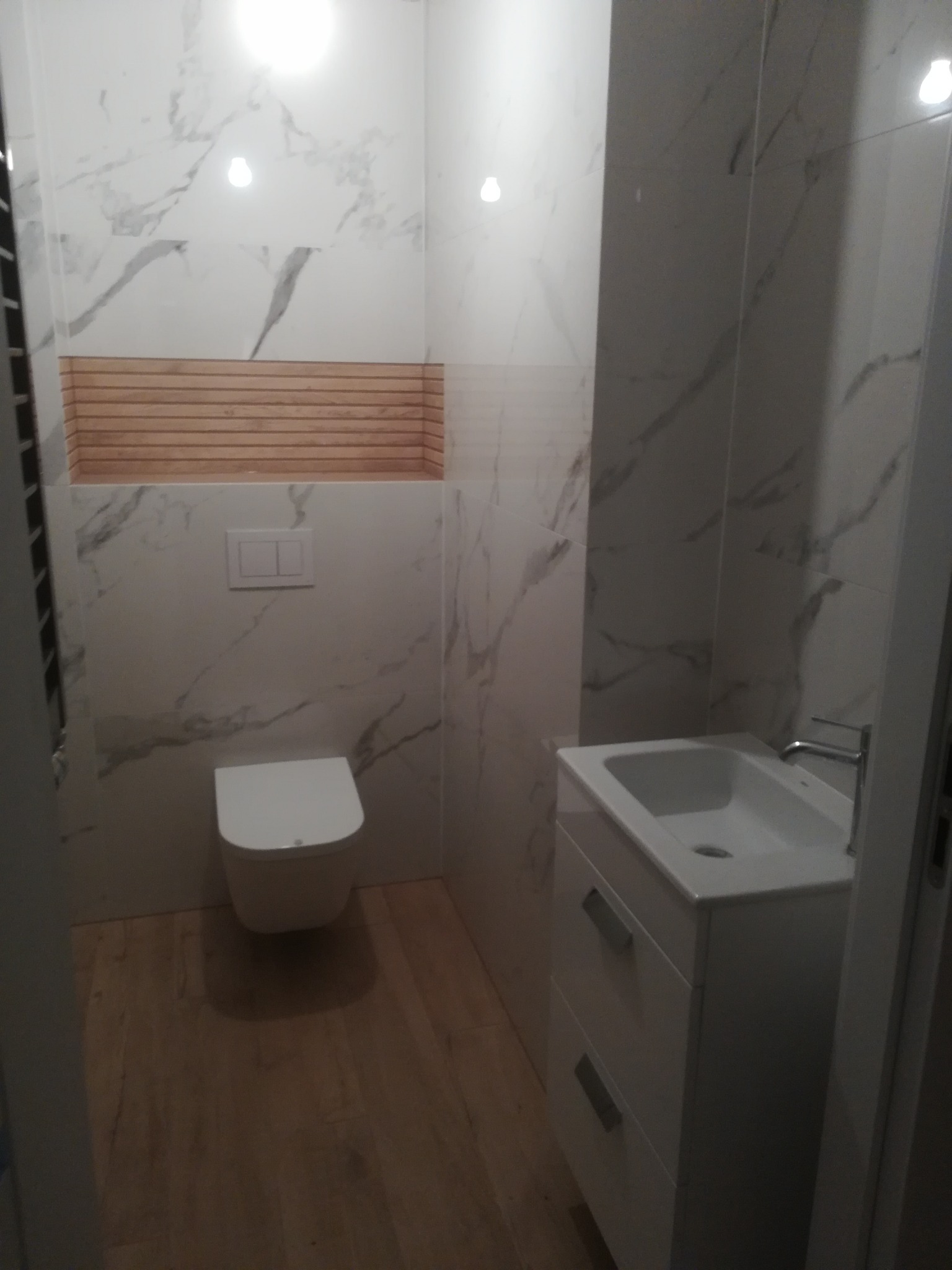 Wykończona łazienka z płytkami imitującymi marmur, drewnianą wstawką nad toaletą, podwieszaną miską WC i białą szafką z umywalką.