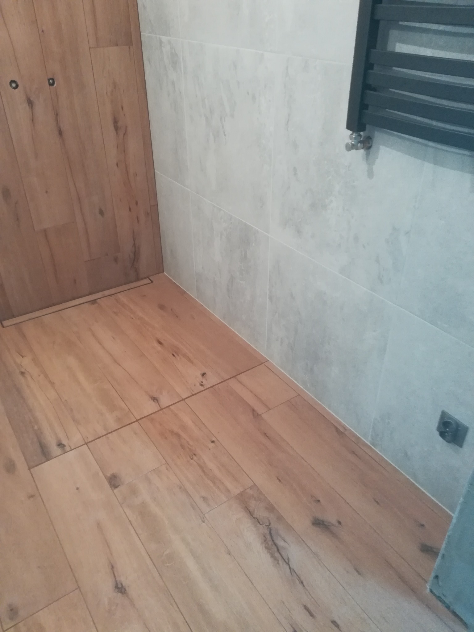 Wykończona łazienka z drewnopodobną podłogą, szarymi płytkami na ścianie i grzejnikiem drabinkowym.