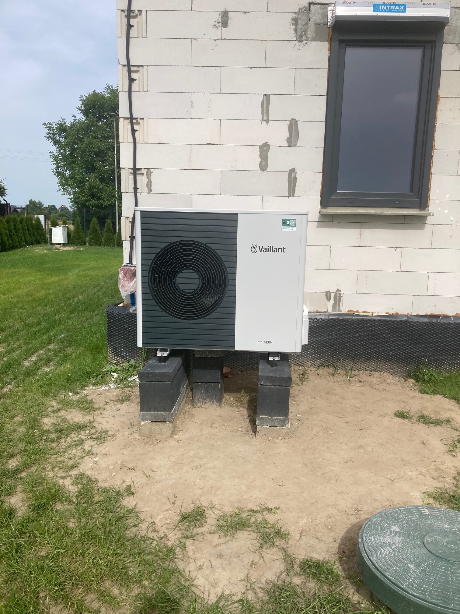 Zewnętrzna jednostka pompy ciepła Vaillant aroTHERM zamontowana na betonowych bloczkach przy ścianie budynku z białego bloczka, widoczne okno z roletą zewnętrzną, trawa i ziemia wokół.