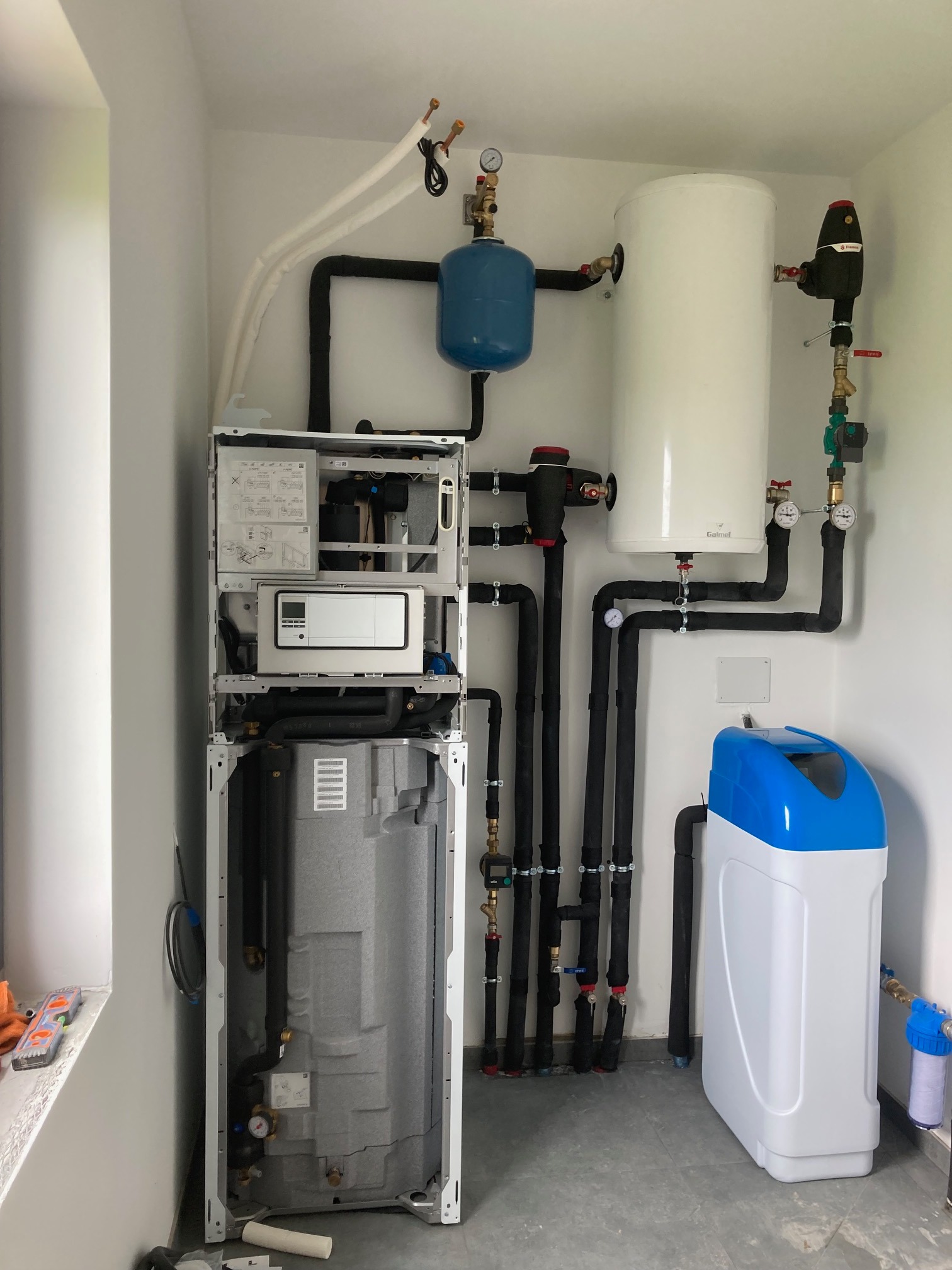 Zestaw urządzeń instalacji grzewczej w pomieszczeniu w Krzyszkowie: pompa ciepła, zbiornik buforowy, zmiękczacz wody, liczne rury i zawory, widoczne narzędzia w tle na parapecie.