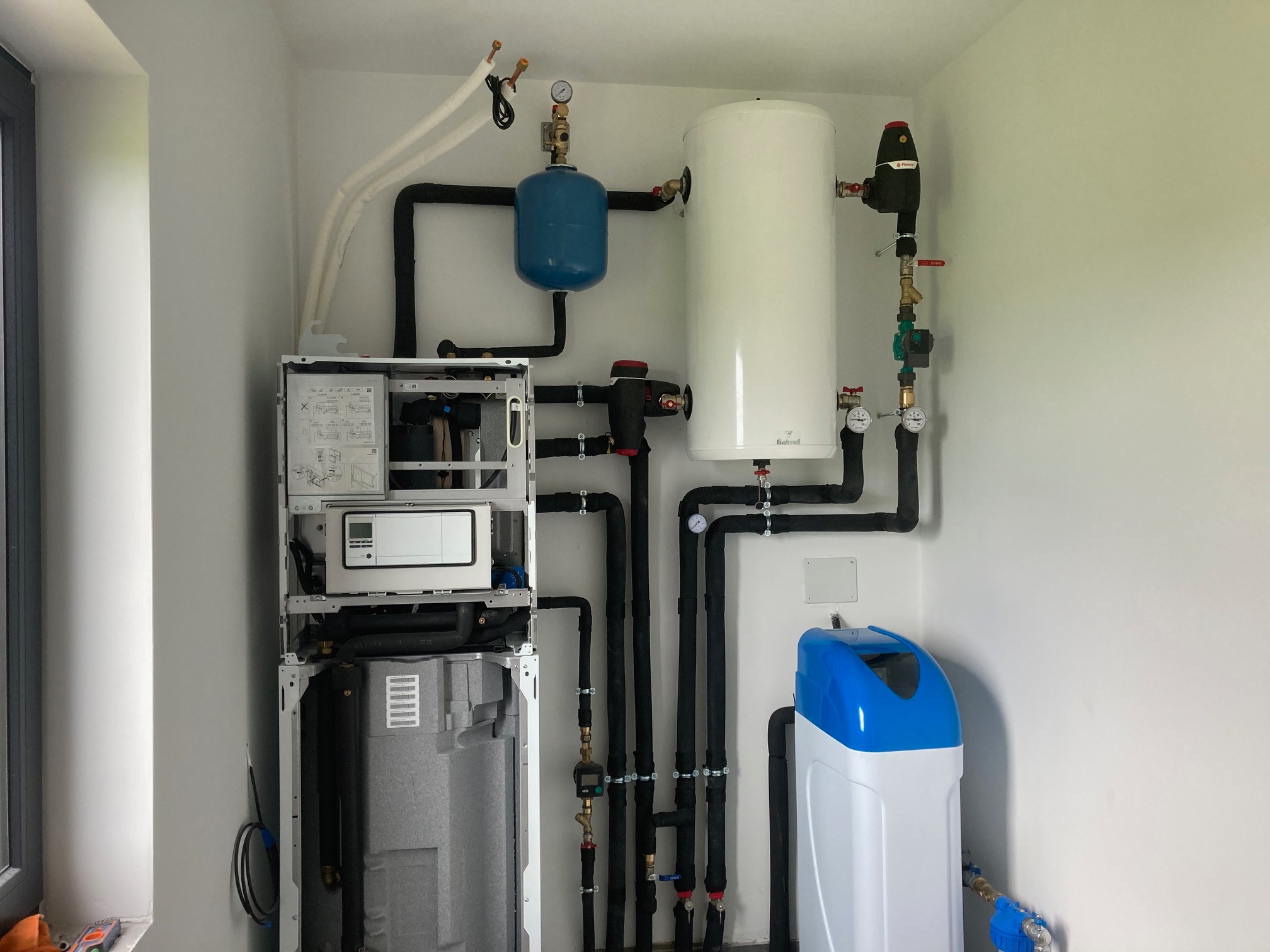 Kompletna instalacja kotłowni z zbiornikiem akumulacyjnym, naczyniem przeponowym i stacją uzdatniania wody, rury w izolacji termicznej prowadzone wzdłuż białych ścian.