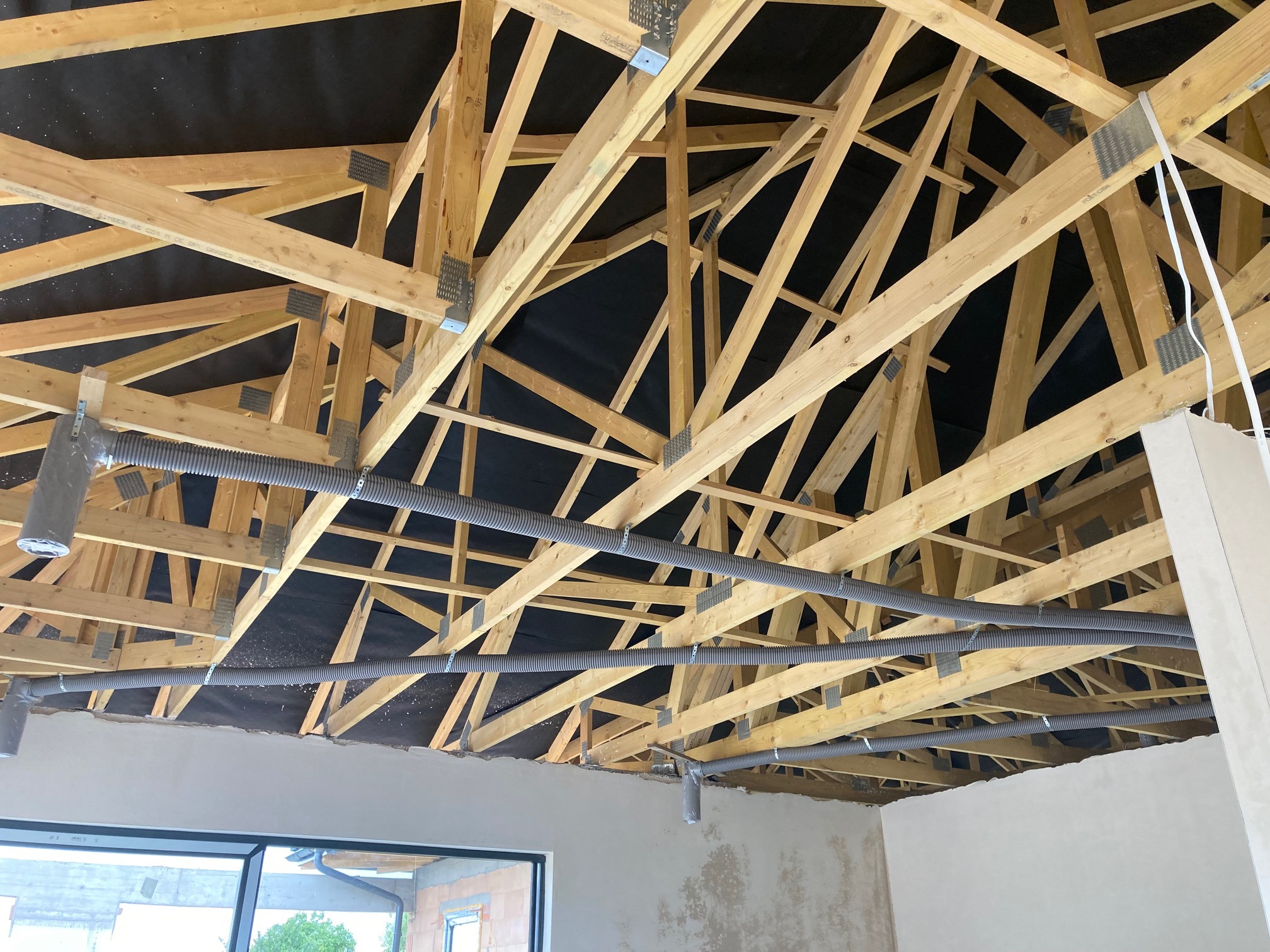 Wnętrze w trakcie budowy z widoczną drewnianą konstrukcją dachu i szarymi, elastycznymi przewodami wentylacyjnymi zamontowanymi pod sufitem, widok z dołu na czarną membranę dachową.