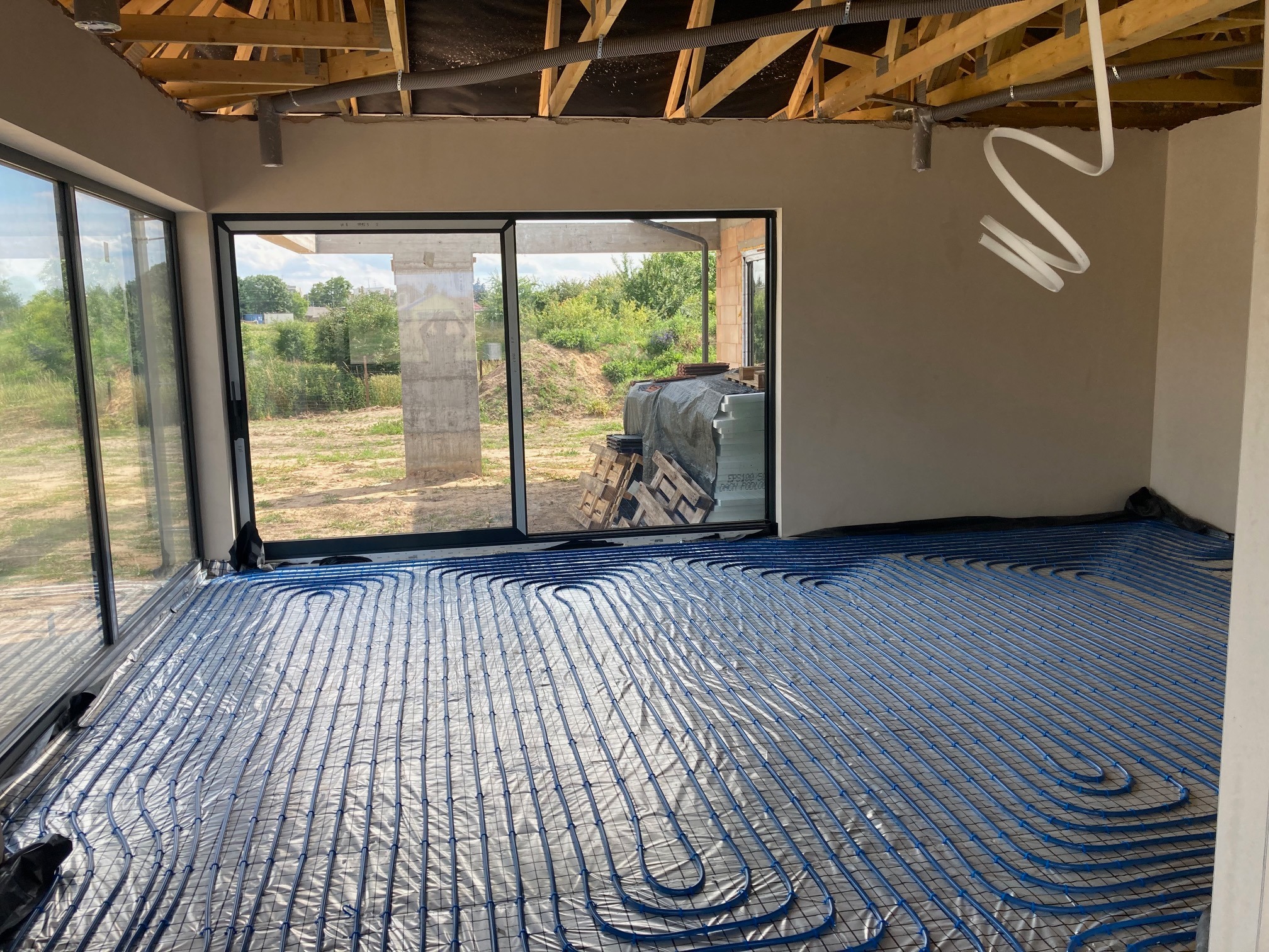 Niebieskie rury instalacji ogrzewania podłogowego ułożone na folii w surowym pomieszczeniu z dużymi oknami, widoczne drewniane elementy konstrukcji dachu i białe rury wentylacyjne pod sufitem.